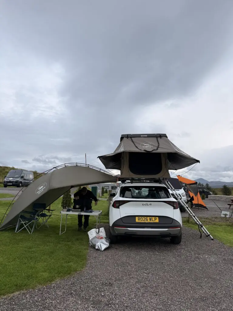 person cooking under awning next to car with rooftop tent